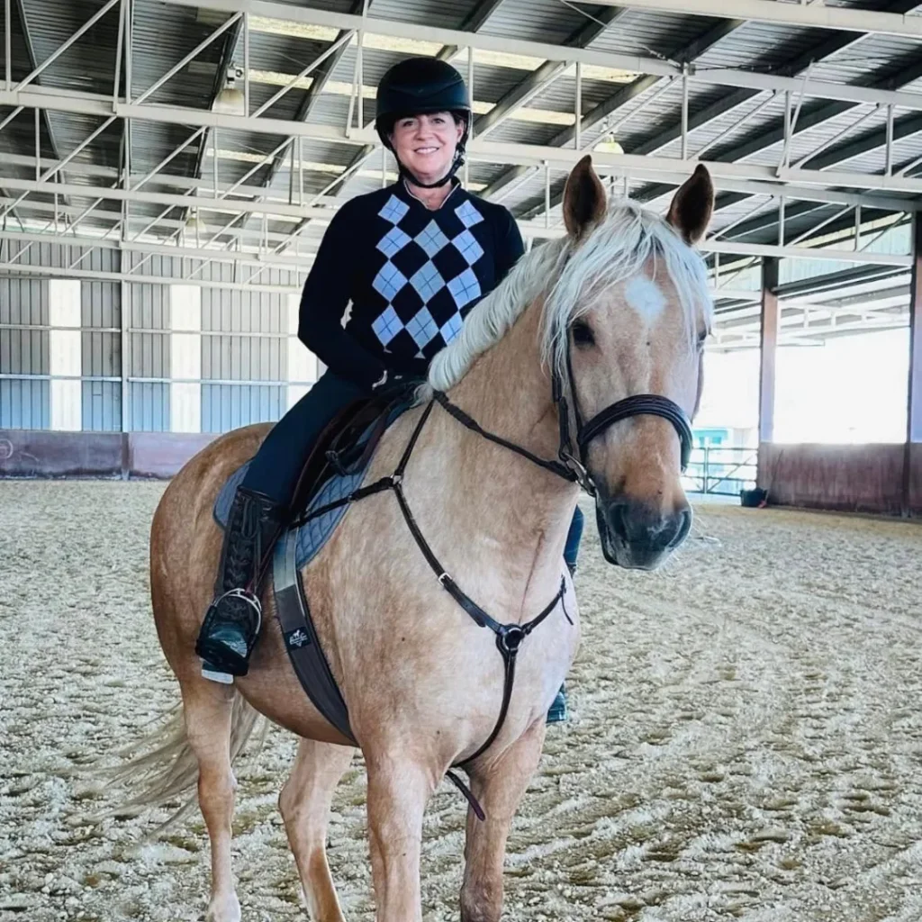 A person wearing a helmet, sweater with a diamond pattern, and riding boots sits smiling on a light brown horse inside an indoor riding arena with a sandy floor and metal roof.