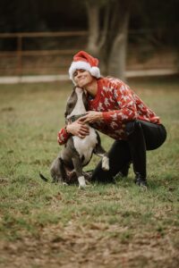 A person wearing a Santa hat and festive red sweater kneels on grass, smiling and hugging a dog that is affectionately nuzzling their face after a fun session of dressage training. Trees and a fence are visible in the blurred background.
