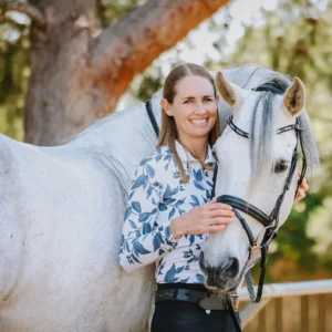 A woman with long brown hair wearing a floral shirt stands beside a white horse, smiling and gently holding its bridle. They are outdoors with trees in the background, showcasing their bond through dedicated dressage training.