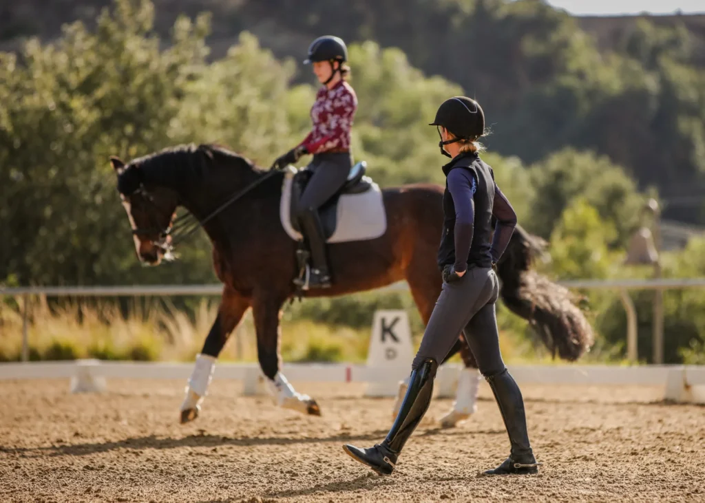 A person on horseback rides in a sandy arena during a workshop, while another in riding gear walks alongside, observing and instructing. Both wear helmets, framed by trees and sunlight in the background.