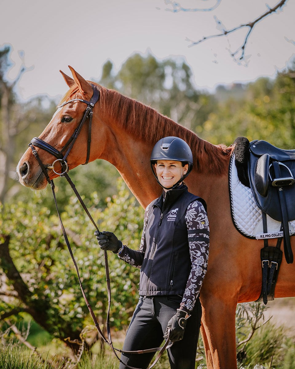 A smiling woman wearing a helmet and riding gear stands beside a chestnut horse with a black bridle and white saddle pad, holding its reins outdoors in a sunlit, green setting.