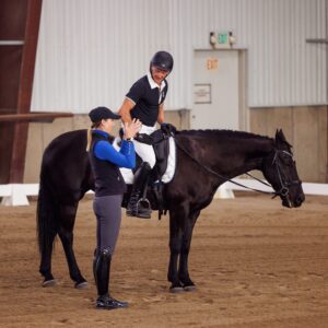 A man in riding gear sits on a black horse, smiling, while a woman in equestrian attire stands beside them inside an indoor riding arena, clapping her hands in encouragement at the Dressage Foundations event. Early Bird spots open for July 2026!.