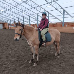 A rider in a helmet and jacket sits on a bay horse inside an indoor riding arena with a dirt floor and high, glass-paneled roof, preparing to move off at a collected walk across the arena.