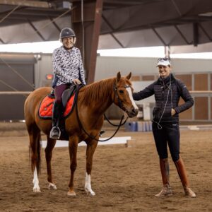 A student sits astride a chestnut horse with a red saddle pad, positioned at halt within an indoor arena. Another woman stands at the horse’s shoulder, smiling warmly while holding the reins, expressing gratitude and camaraderie reminiscent of the supportive atmosphere often found at dedicated dressage clinics or bootcamps.