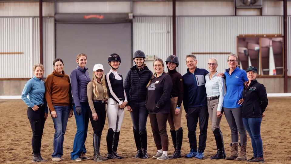 A group of thirteen people, some wearing equestrian helmets and riding attire, stand side by side smiling in an indoor riding arena with sandy flooring and metal walls.