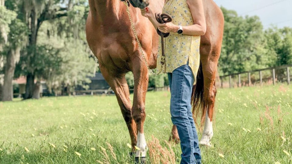 Strides Member Janet, wearing a straw hat, yellow sleeveless shirt, and blue jeans, stands outdoors in a grassy field, holding the reins of a brown horse. Trees and a clear sky form the background.
