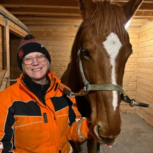 A smiling person in an orange jacket and winter hat stands next to a brown horse with a white heart-shaped marking on its forehead inside a wooden stable.