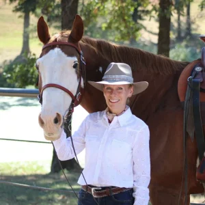 A smiling woman in a white shirt and gray hat stands beside a brown horse with a white face, holding its bridle. Trees and a fenced area are visible in the background.