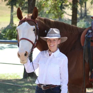 A smiling woman in a white shirt and tan hat stands beside a brown horse with a white face, holding its bridle outdoors on a sunny day. Trees and a fence are visible in the background.