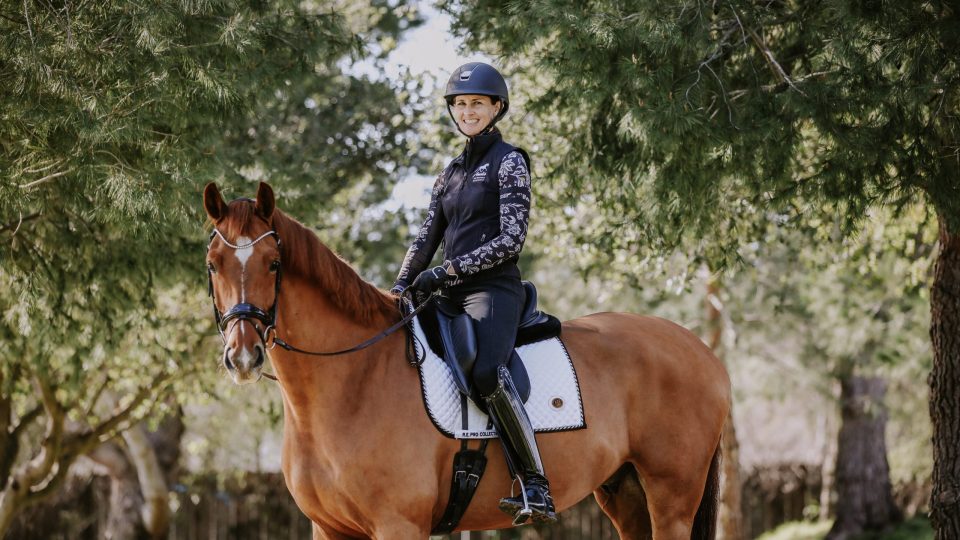 A person wearing a helmet, black riding vest, and boots sits on a brown horse with a white saddle pad, outdoors among green trees and sunlight—a scene reminiscent of August 2023 News highlights.