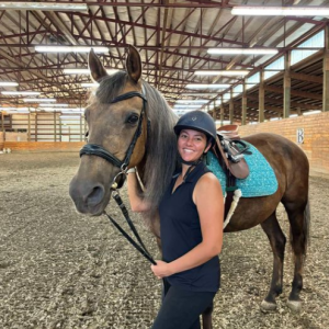 A woman wearing a helmet and black riding attire stands smiling beside a brown horse with a turquoise saddle pad in an indoor riding arena.