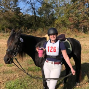 A smiling equestrian in white riding pants and helmet stands outdoors holding a drink, one hand on the reins of a black horse. The rider’s shirt has the number 126. Trees and grass fill the background—perfect after attending a Flying Changes Webinar.