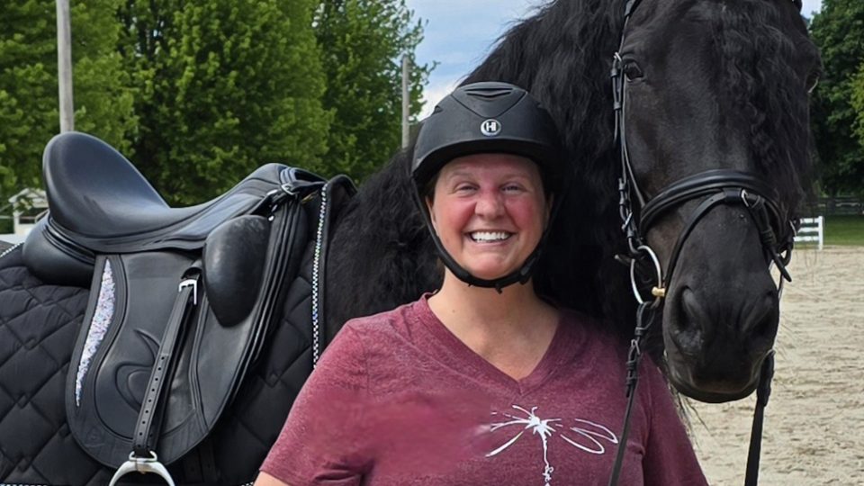 Strides member Kris, smiling in a maroon shirt and black riding helmet, stands beside a black horse with a braided mane on a sandy riding arena, framed by trees in the background.