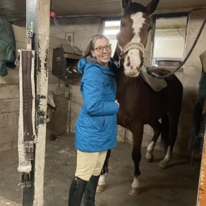 A woman in a blue jacket and riding boots smiles while standing next to a brown horse with a white blaze on its face inside a stable. The horse is wearing a halter and a saddle pad draped over its back.
