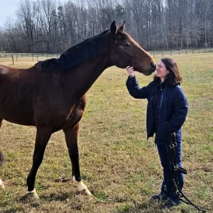 A woman in a dark jacket smiles while gently holding the muzzle of a brown horse in a grassy field with trees in the background.