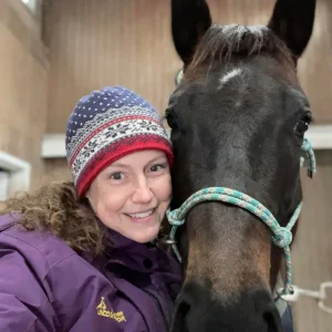 A woman in a purple jacket and colorful winter hat smiles next to a dark brown horse wearing a teal rope halter in a wooden stable.