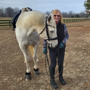 A woman wearing sunglasses and riding gear stands next to a white horse in a fenced outdoor area with dry grass and trees in the background. The horse is saddled and wearing protective boots.