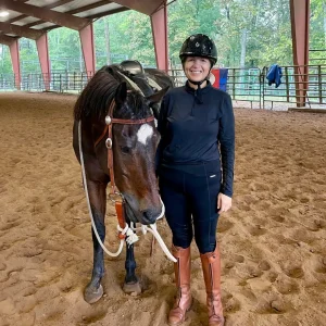 A woman in riding gear and helmet stands smiling beside a brown horse in an indoor riding arena with sandy ground and trees visible outside the structure.