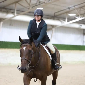 A person wearing a helmet and riding gear smiles while riding a brown horse inside an indoor arena with a sandy floor and a high ceiling.