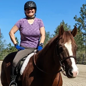 A smiling woman wearing a helmet, purple shirt, and blue gloves is sitting on a brown horse with a white face marking. They are outdoors in a fenced area with trees and a clear blue sky.