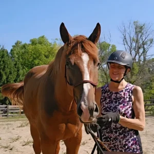 A woman wearing a helmet and gloves stands beside a brown horse, smiling and holding its reins. They are outdoors on a sunny day, with trees and a wooden fence in the background.