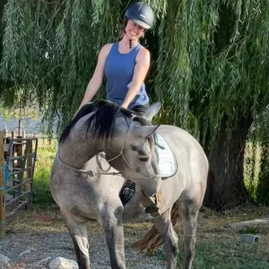 A smiling woman wearing a helmet and blue tank top sits on a gray horse, gently hugging its neck. They are outdoors, with a large leafy tree in the background.