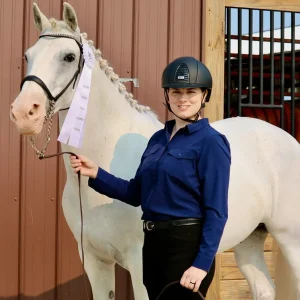 A smiling person in a navy shirt and helmet holds a white ribbon and stands beside a light-colored horse with braided mane outside a stable with brown siding and a wooden door.