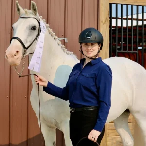 A person in a helmet and blue shirt stands beside a white horse with braided mane, holding a first-place ribbon. They are both in front of a brown barn wall and open stable door.