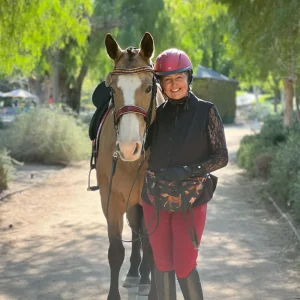 A smiling woman in equestrian gear stands next to a light brown horse with a white face marking on a sunlit dirt path surrounded by green trees.