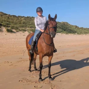 A person wearing a helmet, jeans, and a patterned shirt is sitting and smiling on a brown horse on a sandy beach with grassy dunes in the background under a clear blue sky.