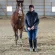 A woman wearing a dark jacket, jeans, and boots stands indoors on dirt beside a brown horse, holding its reins. The background shows wooden beams and metal siding.