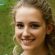 A young woman with wavy blonde hair smiles at the camera. She has blue eyes and is wearing a light top. The blurred green background suggests an outdoor setting, perfect for a fresh 2024 Sale Page or Training Scale promotion.