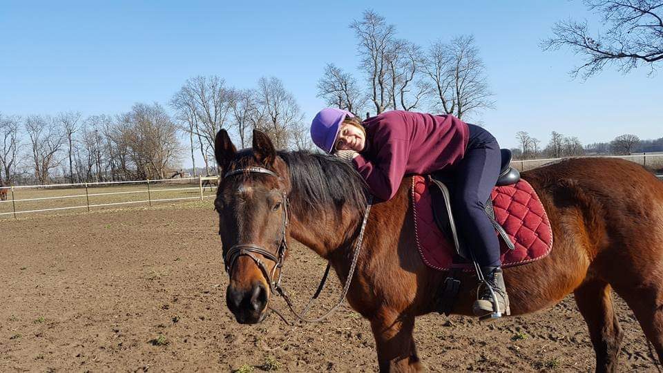 Marcee, a member of Strides, wears a purple helmet and maroon sweater while hugging the neck of a brown horse with a red saddle pad in an open field on a sunny day, surrounded by trees and fences.