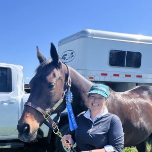 A smiling woman in a green cap and white pants stands next to a brown horse with a blue ribbon, after a 5 Day Riding Bootcamp, with a horse trailer and white truck in the background on a sunny day.