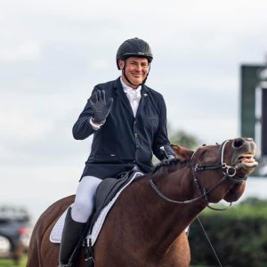 A smiling equestrian in a black helmet and jacket waves while riding a brown horse that is stretching its neck and curling its lips. Blurred greenery and a scoreboard are visible in the background.