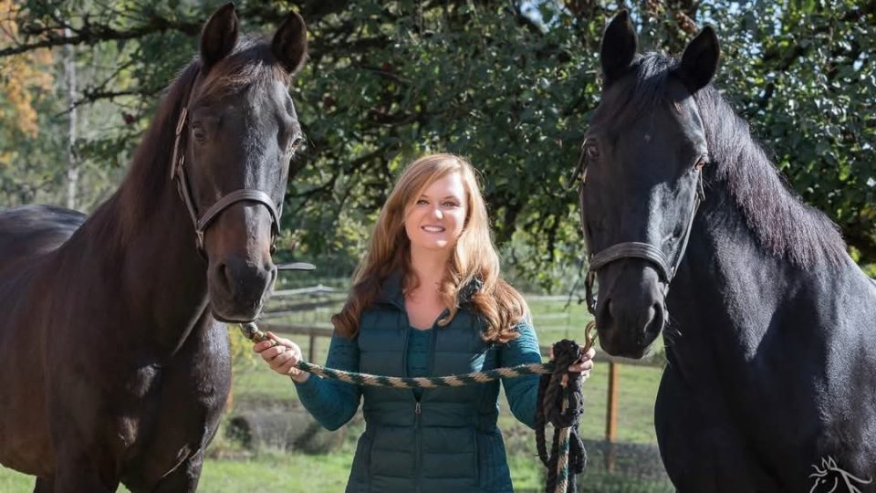 Strides member Shonnessy, a woman with long light brown hair in a teal vest, stands outdoors holding the reins of two large black horses, one on each side, with trees and a fence in the background.