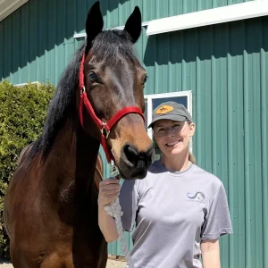 A woman wearing glasses and a cap stands beside a brown horse with a red halter, smiling in front of a green metal building on a sunny day.