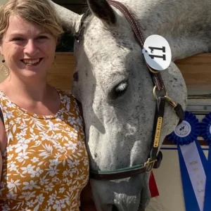 A woman in a yellow floral top smiles beside a gray horse wearing a bridle and number tag 11, with its head leaning affectionately on her. Blue and white ribbons hang in the background.