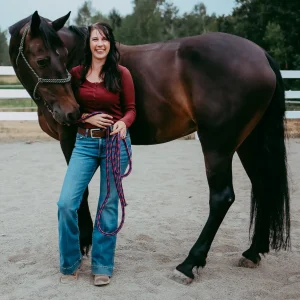 A woman wearing a red long-sleeve shirt and blue jeans stands smiling next to a dark brown horse in an outdoor fenced area, holding a purple and pink lead rope. Trees are visible in the background.