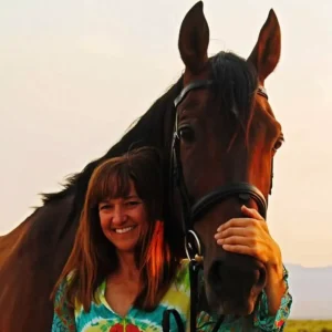 A woman with long brown hair smiles while standing next to a brown horse, resting her hand gently on its face. The background shows a soft, outdoor light.