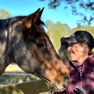 A woman wearing a maroon jacket and black cap smiles and looks at a brown horse with a white stripe on its face. They stand near a wooden fence, with trees and a blue sky in the background.