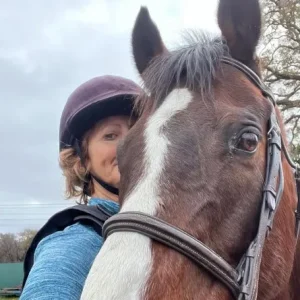 A person wearing a helmet and blue top stands close behind a brown horse with a white blaze on its face, both looking at the camera. The background shows a cloudy sky and some trees.