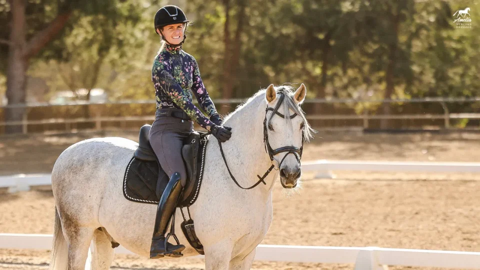 Amelia, a smiling equestrian wearing a helmet and floral jacket, rides a white horse in an outdoor arena on a sunny day, reflecting the spirit of Grassroots Giving with trees and riding barriers in the background.