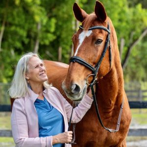 Janet, a Strides member with long blond hair, smiles as she stands outdoors in a light pink jacket and blue shirt, holding the bridle of a tall brown horse with a white blaze. Lush green trees fill the background.