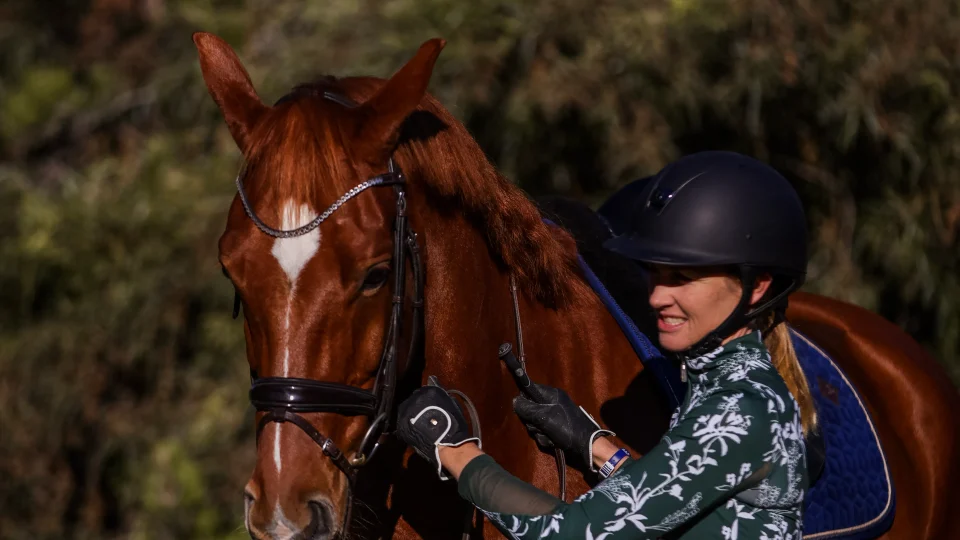 Amelia, in a riding helmet and floral shirt, stands beside a chestnut horse with a blue saddle pad, smiling and holding the reins outdoors with trees in the background—ready to master groundwork and piaffe.
