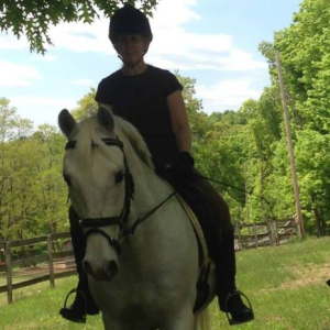 A person wearing a helmet and dark clothing rides a white horse outdoors on a grassy area, with green trees and a wooden fence in the background. Part of a brown dog is visible at the lower right—a perfect scene for the 2025 Riding Bootcamp.