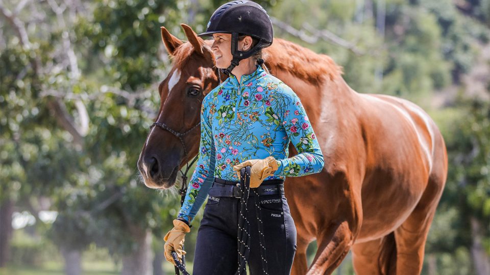 Amelia, wearing a helmet, floral shirt, gloves, and riding pants, leads a chestnut horse by its reins outdoors with trees behind her. Text reads: September News 2023 Newsletter.